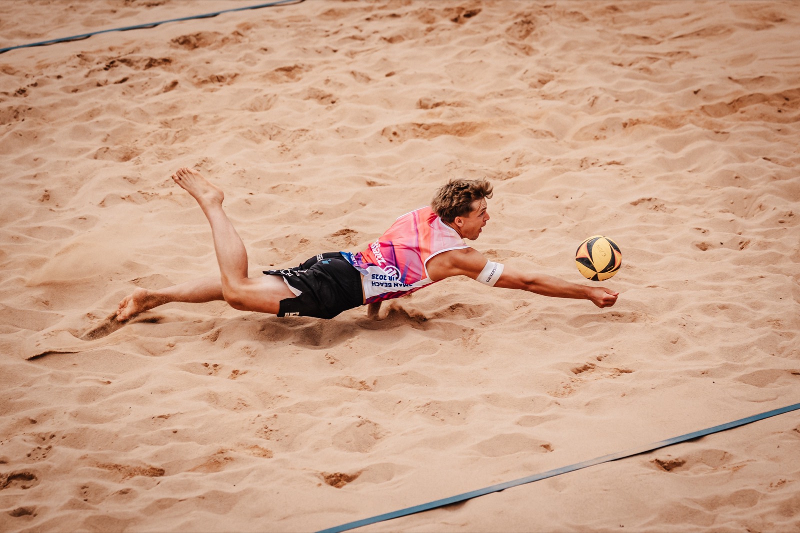 Ein Beachvolleyballer hechtet sich flach über den Sand und streckt den Arm nach dem Ball.